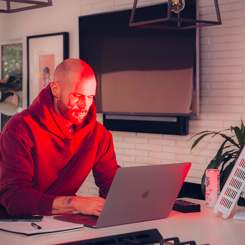 Man in hoodie using a red light teraphy device , laptop at a desk with a TV and decor in the background