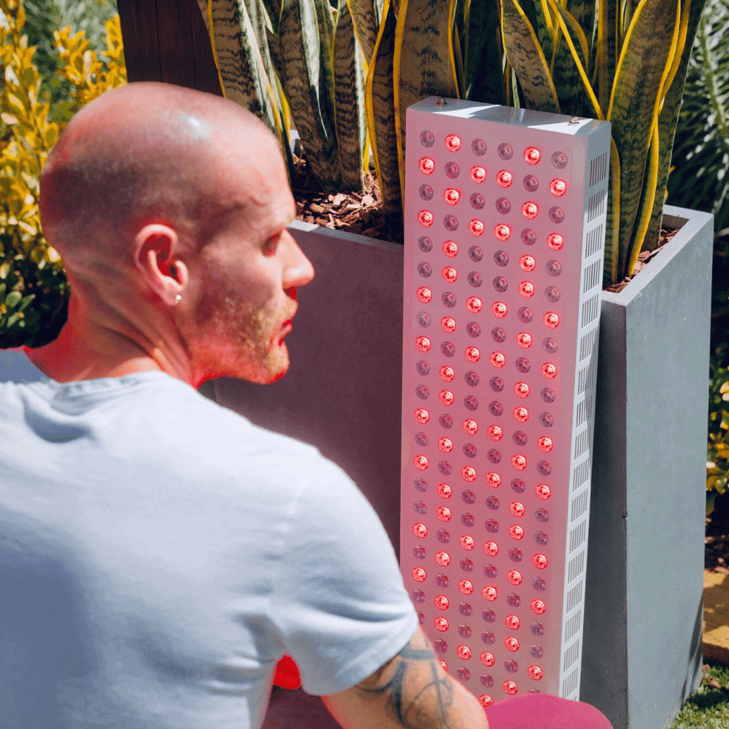 Man sitting next to a tall rectangular red light teraphy panel outdoors.