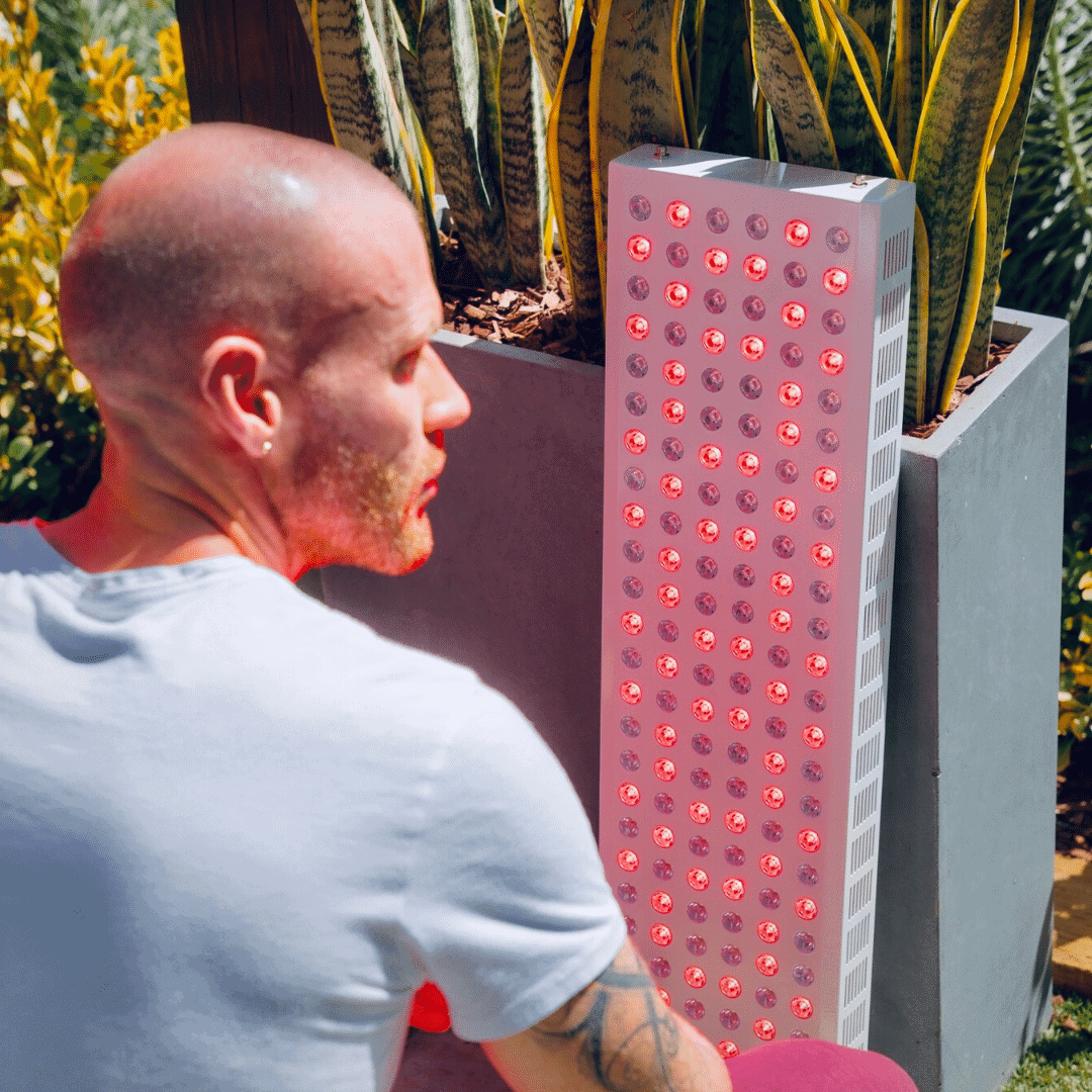 Man sitting next to a tall rectangular red light teraphy panel outdoors.